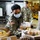 Two U.S. Army soldiers wearing camouflage uniforms and blue surgical masks are working in what appears to be a food bank or distribution warehouse. The foreground soldier, a woman, is sorting and handling packaged baked goods and food items on a table. The setting suggests a military humanitarian or community support operation, likely during the COVID-19 pandemic given the mask usage.