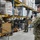 Several military personnel in camouflage uniforms are working inside a large warehouse or food distribution facility. One soldier is filming with a video camera while others handle cardboard boxes and inventory on high shelving racks. The scene appears to document a military logistics or humanitarian aid operation.