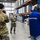 A military service member in OCP (Operational Camouflage Pattern) uniform is being interviewed by a NewsCenter 7 television news crew inside a large warehouse or distribution facility. The scene shows two media crew members — one operating a camera and one holding a boom microphone — along with another uniformed military member photographing the subject. The warehouse is stocked with palletized goods on tall orange and blue industrial shelving racks.