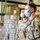 Two U.S. Army soldiers wearing surgical face masks stand inside a warehouse or distribution facility surrounded by stacked cardboard boxes and palletized goods. Both are dressed in OCP (Operational Camouflage Pattern) uniforms with 'U.S. ARMY' tape visible on their chests. The setting suggests a logistics, food bank, or emergency relief support operation.