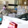A U.S. Army soldier wearing camouflage uniform, a patriotic face mask, and blue latex gloves is directing or assisting at what appears to be a food distribution event. The soldier is standing near a white vehicle's trunk in a warehouse setting surrounded by pallets of canned goods and boxes labeled 'Fresh Vegetables'. A sign in the background reads 'AO CSFP FEB', suggesting this is a Commodity Supplemental Food Program (CSFP) distribution operation.