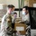 Three U.S. Army soldiers in OCP (Operational Camouflage Pattern) uniforms and face masks are working in a warehouse, handling boxes of food supplies. Two soldiers in the foreground are transferring a cardboard box labeled 'KEEP FROZEN' into or out of a vehicle. The setting appears to be a food distribution operation, likely during the COVID-19 pandemic based on the masks worn.
