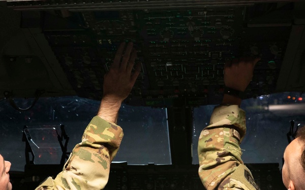 Two military personnel in camouflage uniforms (OCP/MultiCam pattern) are seated in what appears to be a large military aircraft cockpit, likely a C-17 Globemaster or similar transport aircraft. Both individuals are reaching upward to interact with the overhead control panel, suggesting pre-flight checks or systems configuration. The cockpit displays multiple digital screens and instrument panels illuminated with green and blue lighting.