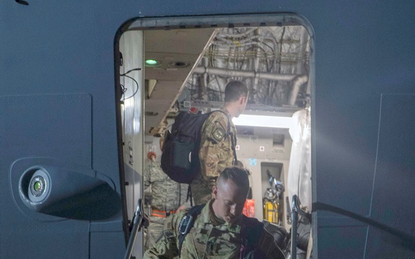 Military personnel in camouflage uniforms are deplaning from a military aircraft via a stairway at night. The foreground soldier is carrying a maroon bag labeled 'Collierville Dragons Track &amp; Field' along with a tactical backpack. At least two additional soldiers are visible inside the aircraft doorway.