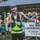 A tattooed male runner wearing bib number 135 poses near the finish area of what appears to be a trail ultramarathon in Hawaii. He is wearing a hydration vest, headlamp, and neon yellow running gear, giving a thumbs-up gesture. A bilingual sign in Hawaiian and English reads 'Aole Makou E Ho'ohikiwale Kela / We Wouldn't Want It To Be Easy.'