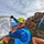 A person dressed in athletic outdoor gear, including a bright blue long-sleeve shirt, yellow cap branded 'OS1st', orange face gaiter, and reflective sunglasses, holds up a white flag or banner at what appears to be a volcanic mountain summit. The banner contains partial text reading 'UNITED' and a handwritten inscription referencing 'Pearl Harbor Memorial July 2012'. Rocky volcanic terrain and a cloudy sky form the backdrop, with at least one other person visible in the background.