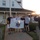 A group of five men pose for a photo while holding a United States Coast Guard flag with the motto 'Semper Paratus' and the year '1790'. They are standing on a brick walkway in front of a white clapboard building that appears to be a Coast Guard museum or station. A sixth person on the right side of the frame is photographing the group during what appears to be sunset or sunrise.