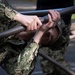 A U.S. Navy service member in NWU Type III (woodland digital camouflage) uniform is performing a pull-up or bar hang exercise on a metal horizontal bar. The individual is gripping the bar tightly with both hands, with their chin near or at bar level, suggesting a physical fitness or obstacle course activity. A 'U.S. NAVY' patch and partial 'USN' tape are visible on the uniform.