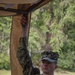 A U.S. Navy service member wearing NWU (Naval Working Uniform) Type III camouflage is holding up a tan/coyote-colored canopy or shelter structure with one raised arm. The name tape reads 'WOODARD' and the branch tape reads 'U.S. NAVY.' The setting appears to be an outdoor wooded or park-like environment.