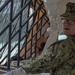A U.S. Navy service member in NWU Type III (green digital camouflage) uniform and cover is handling a metal frame structure, likely assembling or positioning equipment. The setting appears to be a temporary or field environment with plastic sheeting and tent-like structures visible in the background. The individual is wearing glasses and appears to be speaking or directing while working.