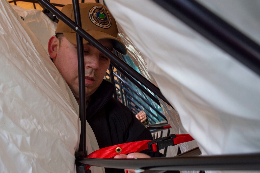 A person wearing a tan baseball cap with an emblem is working inside or around a structure with black metal framing and white plastic sheeting. They appear to be handling a red strap or harness with a black buckle/clasp. The image is taken from a close, low angle through the frame structure.