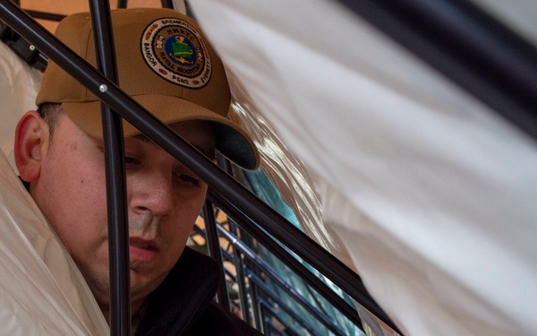 A person wearing a tan baseball cap with an emblem is working inside or around a structure with black metal framing and white plastic sheeting. They appear to be handling a red strap or harness with a black buckle/clasp. The image is taken from a close, low angle through the frame structure.
