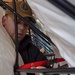 A person wearing a tan baseball cap with an emblem is working inside or around a structure with black metal framing and white plastic sheeting. They appear to be handling a red strap or harness with a black buckle/clasp. The image is taken from a close, low angle through the frame structure.