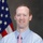 A formal portrait of a middle-aged man with short reddish-brown hair, freckles, and a slight smile. He is dressed in a light blue checkered dress shirt and a pink tie with a small repeating pattern. An American flag is partially visible in the background, suggesting an official or government-related setting.