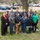 A group of approximately eleven people pose for a group photo outdoors on a grassy area near a black iron fence and brick pillars. Several individuals are wearing jackets or sweatshirts with what appears to be university or organizational branding. A large decorative anchor prop is visible on the right side of the group.