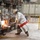 Two industrial workers in full heat-protective gear are operating near a high-temperature furnace in what appears to be a metal smelting or foundry facility. They are using long-handled tools to interact with molten or extremely hot material visible as glowing orange at the furnace opening. The setting is a large industrial plant with concrete floors, yellow safety railings, and heavy machinery.