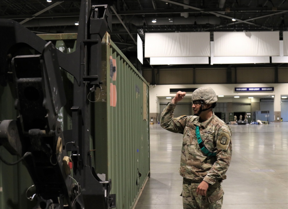 A U.S. Army soldier in full combat uniform and helmet is directing or signaling near a large military forklift and green shipping container inside what appears to be a convention center or arena. The soldier is giving a hand signal with a raised fist, likely communicating with equipment operators. The background shows 'Bud Light' signage, confirming the indoor venue is a commercial event space being used for military operations.
