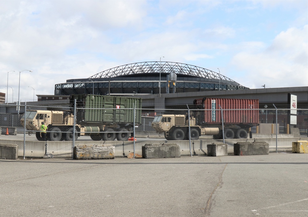 The image shows a secured port or industrial facility with military vehicles staged behind a chain-link fence reinforced with concrete barriers. In the background, a large domed stadium with a distinctive arched roof structure is visible, consistent with a retractable-roof sports venue. A sign marked '46' is visible on the right side, suggesting a numbered gate or pier.
