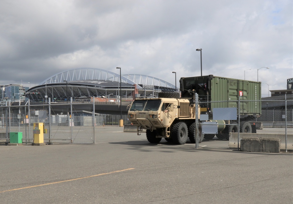 A military tactical truck, appearing to be a tan/desert-colored LMTV or FMTV, is positioned in a secured parking area near what appears to be Lumen Field (formerly CenturyLink Field) in Seattle, Washington. The vehicle is hauling a large olive-drab military cargo container. The area is enclosed by chain-link fencing with a gate, suggesting a controlled or restricted zone.