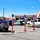 A parking lot outside an Army &amp; Air Force Exchange Service (AAFES) store, featuring a curbside pickup lane marked with numbered poles (1-4) on red bases labeled 'PICKUP HERE'. Multiple vehicles are parked in the lot, including a red Ford pickup truck prominently in the foreground. The scene appears to be a military base exchange store with curbside pickup infrastructure set up, likely implemented during or after the COVID-19 pandemic period.