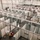 An elevated view of a large warehouse or convention center being converted into a temporary medical facility or field hospital. The space is filled with rows of modular white-walled cubicles with privacy curtains, connected by overhead piping infrastructure. Workers in high-visibility safety vests are actively installing equipment and completing construction.