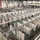 A large convention center or warehouse facility is being converted into a temporary medical or emergency field hospital, with hundreds of individual patient cubicles being constructed using white partition walls and gray privacy curtains. Workers in high-visibility safety vests are actively installing infrastructure including overhead piping systems (likely oxygen, medical gas, or electrical conduit) running along the ceiling and between rows. The scale of the operation is massive, filling the entire floor of what appears to be an exhibition hall.
