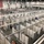 A large convention center or warehouse facility is being converted into a temporary medical or emergency shelter, with hundreds of individual cubicles constructed from white partition walls and curtains. Workers in high-visibility safety vests and hard hats are actively installing infrastructure including overhead piping and electrical conduit. An American flag is visible on the far wall, and a 'Restrooms' sign is posted near the entrance.