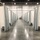 A large convention center or warehouse space has been converted into a temporary medical or quarantine facility, with rows of gray privacy curtains suspended from ceiling-mounted tracks creating individual partitioned bays on both sides of a central corridor. The polished concrete floor reflects overhead lighting, and the space appears to be in an operational or near-operational state. Two workers wearing high-visibility vests and face masks are visible at the far end of the corridor near a large roll-up door.