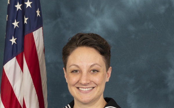 A U.S. Navy enlisted service member poses for an official military portrait in dress blue uniform. The subject is seated at a blue-draped table with an American flag visible in the background. The uniform displays a Petty Officer First Class rating badge, multiple service ribbons, a warfare qualification device, and service stripes.