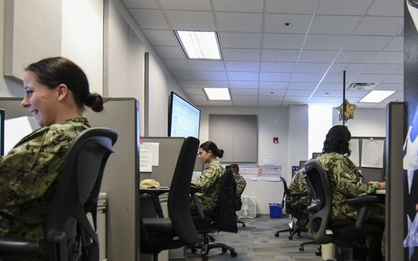 Military personnel in camouflage uniforms are working at computer workstations in an office environment identified as MNCC (MyNavy Career Center). The workspace consists of cubicle-style desks arranged in rows with multiple monitors visible. At least four service members are visible, all appearing to be engaged in computer-based work tasks.