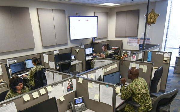 A military office environment showing multiple personnel in Navy Working Uniform (NWU) camouflage pattern seated at cubicle workstations. The open-plan office contains numerous computer monitors, phones, and paperwork-covered cubicle walls. A large display screen is mounted on the wall at the front of the room.