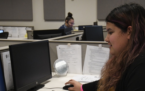 A woman with long curly hair with pink highlights is working at a cubicle workstation in an open office environment. She is using a dual-monitor HP computer setup with a keyboard and mouse, with printed documents clipped to a partition behind her. A second employee is visible in the background at another workstation.