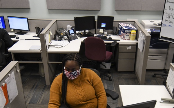 An office environment with cubicle workstations where employees are working at desktop computers. A woman in the foreground wearing an American flag-patterned face mask and orange sweater is prominently visible at her desk. The setting appears to be during the COVID-19 pandemic period, evidenced by mask-wearing and visible social distancing signage on a whiteboard in the background.