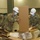 Three U.S. Army soldiers in camouflage uniforms, white hard hats, and blue surgical masks are working inside a large indoor facility, unpacking and assembling equipment from cardboard boxes. The setting appears to be a convention center or large public space being converted for emergency or medical use. The presence of masks and the large-scale setup suggest this is a COVID-19 pandemic response operation.