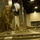 A U.S. Army soldier in OCP (Operational Camouflage Pattern) uniform, wearing a white hard hat and blue surgical mask, is assembling or inspecting military-style cots/beds inside a large convention center or arena. Additional soldiers and workers in hard hats are visible in the background amid stacked equipment and supplies. This appears to document the setup of an emergency field hospital or alternate care facility, likely during the COVID-19 pandemic.
