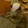 A U.S. Army soldier in OCP (Operational Camouflage Pattern) uniform is kneeling on the floor assembling or adjusting a medical cot or gurney. The soldier is wearing a white hard hat and a blue surgical mask, suggesting this is likely a field hospital or emergency medical facility setup operation. Cardboard packaging debris visible in the background indicates recent unpacking of equipment.