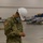 A U.S. Army soldier named Ramirez, wearing OCP camouflage uniform, a white hard hat, and a blue surgical face mask, stands in a large warehouse or convention center facility. The soldier appears to be working near metal bed frames or cot assemblies, likely involved in setting up a field medical or emergency facility. The background shows numerous additional cots or medical equipment staged in the large open space.
