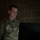 A U.S. Air Force enlisted member in OCP (Operational Camouflage Pattern) uniform is seated at a computer workstation in a dimly lit room with a concrete block wall background. The individual appears focused on the monitor in front of them, illuminated by the screen's glow. Visible uniform markings indicate U.S. Air Force branch affiliation and a Staff Sergeant rank insignia.