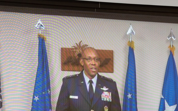 A high-ranking U.S. Air Force officer in dress uniform is displayed on a projection screen, appearing to deliver a video address or presentation. The officer is surrounded by multiple flags including the U.S. flag and Air Force command flags. The image is being projected onto a large screen, visible from the slight scan lines and projection quality.