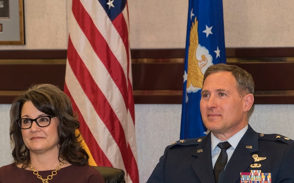 A U.S. Air Force officer in dress uniform sits alongside a civilian woman at what appears to be an official military ceremony or formal event. The officer displays two stars on his shoulders indicating the rank of Major General, along with multiple service ribbons and badges. The setting includes an American flag and U.S. Air Force flag in the background, suggesting a formal military function such as a promotion or change of command ceremony.
