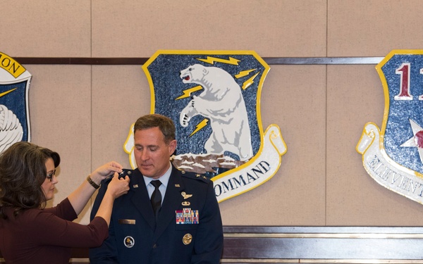 A military promotion or decoration ceremony is taking place in an official conference room. A woman in civilian attire is pinning a new rank insignia or badge onto the shoulder or collar of a U.S. Air Force officer in dress blues. Three military unit emblems are displayed on the wall behind them, identifying the location as associated with Alaskan NORAD Region, Alaskan Command, and Eleventh Air Force.