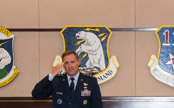 A U.S. Air Force officer in dress blues is rendering a salute while standing in front of three military unit emblems on a wall. The setting appears to be an official military facility associated with Alaskan Command, NORAD, and Eleventh Air Force. The scene suggests a formal ceremony or official photograph.