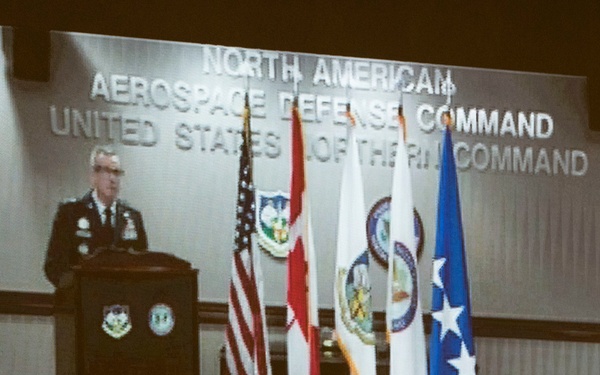 A projected screen display shows a military officer in uniform speaking at a podium during an official briefing or press conference at NORAD (North American Aerospace Defense Command) and United States Northern Command headquarters. The image appears to be a video teleconference or projected presentation being viewed from a conference room. Multiple flags and official insignia are visible behind the speaker.