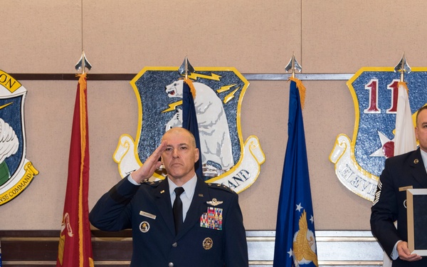 A formal U.S. Air Force military ceremony is taking place in front of organizational insignia and flags. One officer in dress blues is rendering a salute while a second uniformed individual holds a framed medal or award. The setting appears to be an official ceremony room at an Alaskan Command installation, likely a decoration or award presentation.