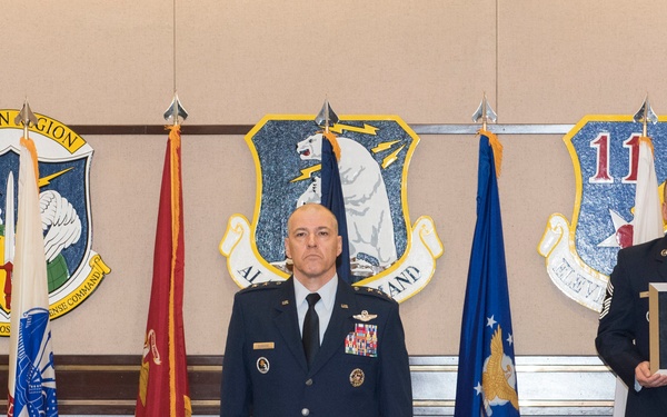 A formal military ceremony is taking place in what appears to be an official U.S. Air Force command room in Alaska. Two uniformed military personnel stand at attention before multiple organizational flags and unit crests, with one individual holding a framed award or certificate. The setting includes insignia from Alaskan Command, North American Aerospace Defense Command (NORAD), and the 11th Air Force.