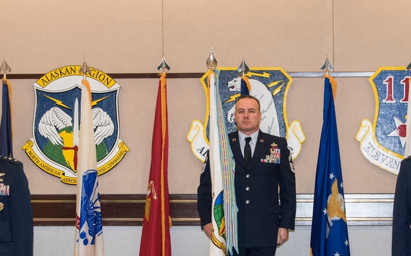 Three U.S. Air Force personnel stand at attention in a formal military ceremony room, flanked by multiple military flags and unit insignia plaques on the wall. The setting appears to be an official ceremony, likely a change of command or award presentation, at an Alaskan Region NORAD installation. A conference table with a camera on a tripod is visible in the foreground, suggesting the event was being recorded or broadcast.