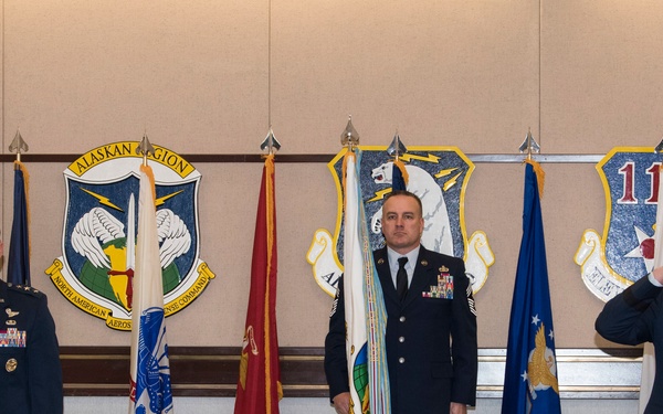 Three U.S. Air Force personnel are standing in a formal military ceremony room, likely at an Alaskan NORAD/11th Air Force installation. Two officers in service dress uniforms flank a senior enlisted member, with one officer rendering a salute. Multiple military flags and unit insignia plaques are displayed on the wall behind them.