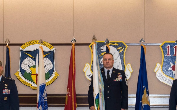 Three U.S. Air Force personnel are standing in a formal military ceremony room, with one officer on the left rendering a salute. The setting features multiple military flags and two unit emblems on the wall, including the Alaskan Region/NORAD shield and what appears to be the 11th Air Force emblem. A camera on a tripod in the foreground suggests the ceremony is being recorded or broadcast.