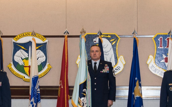 Three U.S. Air Force personnel stand at attention in a formal military ceremony room. The setting features multiple military unit insignia plaques on the wall, including the Alaskan Region and North American Aerospace Defense Command (NORAD), along with numerous military flags. Two officers with rank insignia flank a non-commissioned officer in the center, suggesting a formal command ceremony.