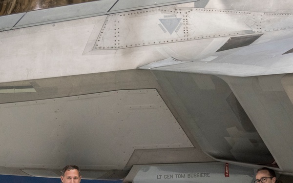 Two U.S. Air Force personnel are crouching beneath a stealth fighter aircraft, likely an F-22 Raptor, inside a hangar. The person on the left, wearing a flight suit, is pointing to a name placard on what appears to be a weapon or external store that reads 'LT GEN TOM BUSSIERE'. The scene appears to be a ceremonial or commemorative photo opportunity.
